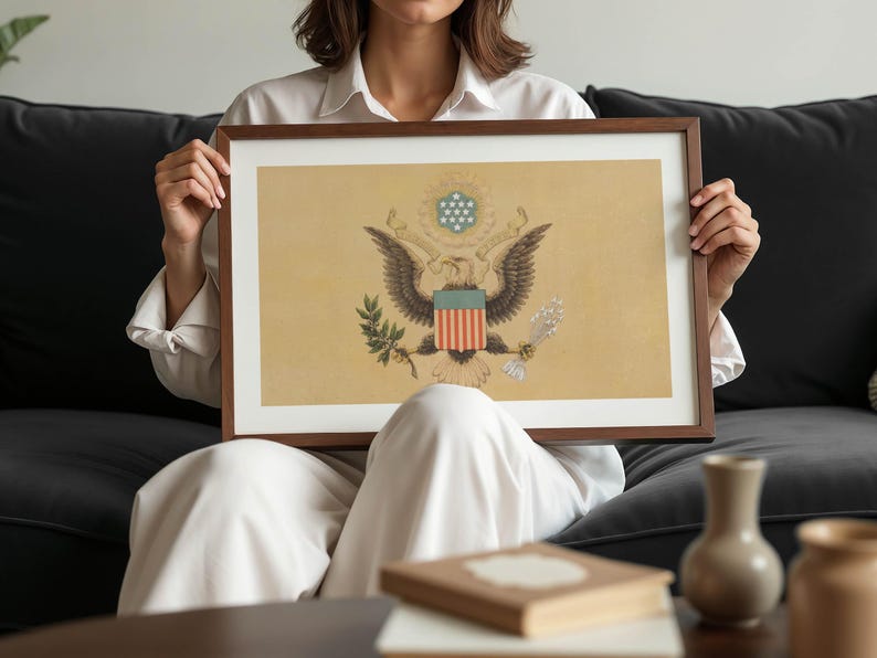 K&ouml;nnte beinhalten: Ein gerahmter Druck mit dem Gro&szlig;en Siegel der Vereinigten Staaten. Das Kunstwerk zeigt den amerikanischen Wei&szlig;kopfseeadler, das Schild und andere Symbole auf beigem Hintergrund. Der Rahmen ist dunkelbraun. Der Druck ist etwa 60 cm breit.