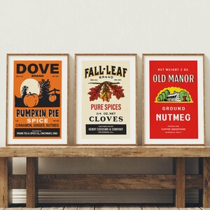 May include: Three framed vintage spice advertisements. The first is orange with a pumpkin illustration, the second is beige with fall leaves, and the third is red with a house illustration. All are displayed on a rustic wooden bench.