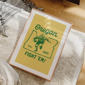 K&ouml;nnte beinhalten: Gerahmtes Poster mit gelbem Hintergrund und gr&uuml;nem Text und Grafiken. Der Text lautet "Oregon" &uuml;ber einer Grafik eines Footballspielers innerhalb der Umrisse des Staates. Unter der Grafik stehen die Worte "FIGHT 'EM!" und das Jahr "1894".