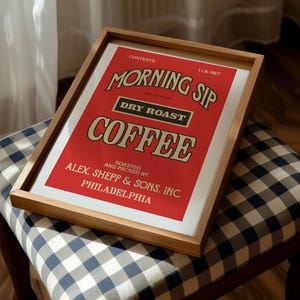 May include: A framed print with a red background, displaying the words "MORNING SIP COFFEE" in a vintage style. The print also includes the text "DRY ROAST" and "ALEX. SHEPP & SONS, INC PHILADELPHIA". The frame is made of light-coloured wood.