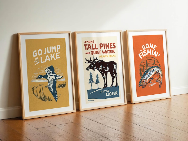 May include: Three framed prints with vintage-style illustrations. The first print features a yellow background with a blue duck in flight and the text "Go Jump in the Lake". The second print features a blue background with a moose and the text "Among Tall Pines and Quiet Water, Heaven Seems a Little Closer". The third print features an orange background with a fish and the text "Gone Fishin'".
