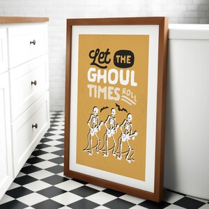 May include: Framed art print with a brown wooden frame. The print has a mustard yellow background with the text "Let the Ghoul Times Roll" in white and black. Three skeleton figures play guitars.