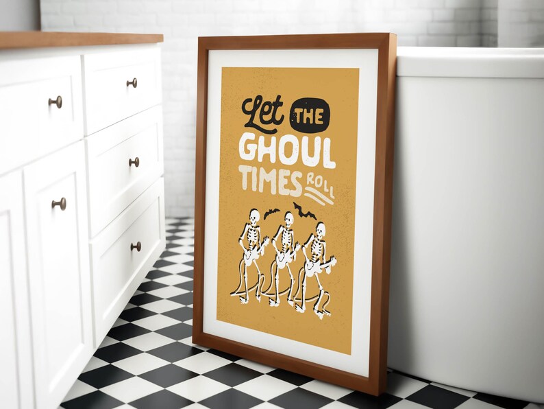 May include: Framed art print with a brown wooden frame. The print has a mustard yellow background with the text "Let the Ghoul Times Roll" in white and black. Three skeleton figures play guitars.