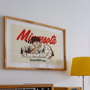 May include: A framed vintage poster with a cartoon illustration of a squirrel paddling a canoe. The poster is in a wooden frame and has the text "Minnesota Land of 10,000 Lakes".