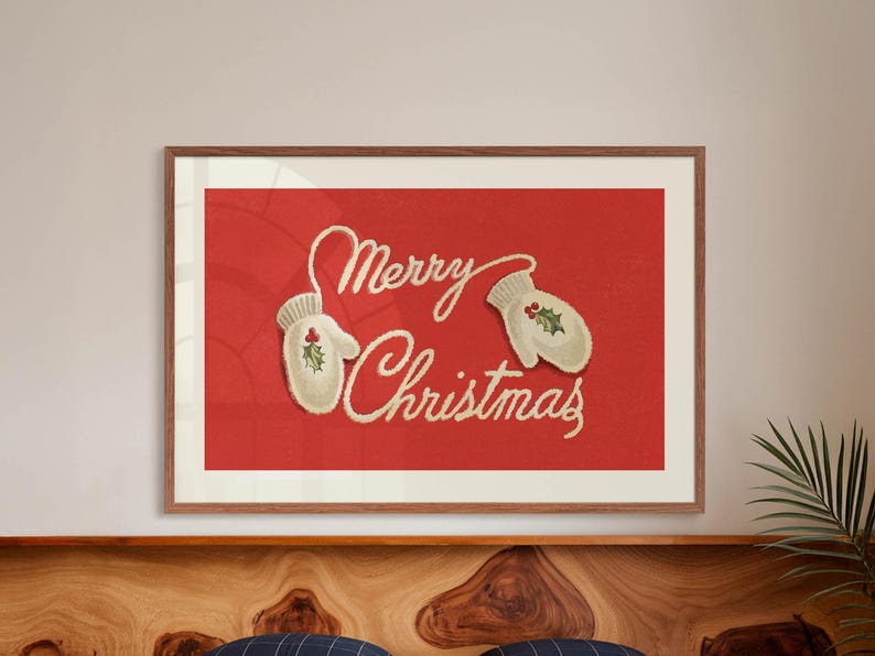 May include: A framed print with a red background and the words "Merry Christmas" in a cursive font. Two off-white mittens with holly accents are on the red background. The frame is a warm brown color.