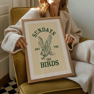 May include: A framed art print with a light beige background and a green eagle illustration. The text "SUNDAYS ARE FOR THE BIRDS" is printed above and below the eagle. The frame is made of light brown wood.