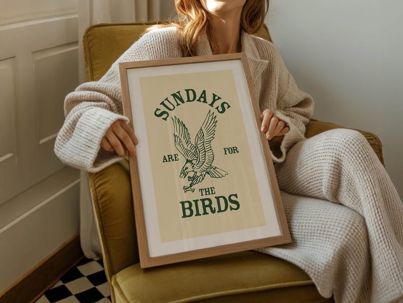 May include: A framed art print with a light beige background and a green eagle illustration. The text "SUNDAYS ARE FOR THE BIRDS" is printed above and below the eagle. The frame is made of light brown wood.