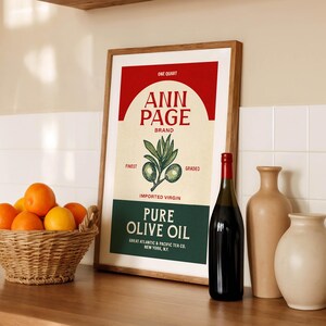 May include: A framed vintage-style poster for "Ann Page" brand pure olive oil, with a red and cream color scheme. A bottle of dark liquid, two beige vases, and a wicker basket of oranges are also present.