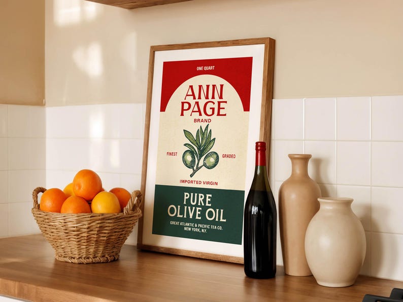 May include: A framed vintage-style poster for "Ann Page" brand pure olive oil, with a red and cream color scheme. A bottle of dark liquid, two beige vases, and a wicker basket of oranges are also present.