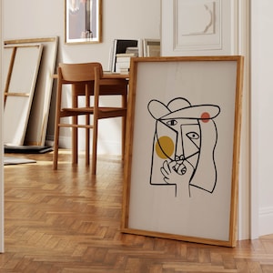May include: A framed abstract line art print of a person's face with a hat, yellow and red circles, and black lines. The print is on a white background and has a wooden frame.