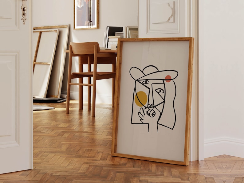 May include: A framed abstract line art print of a person's face with a hat, yellow and red circles, and black lines. The print is on a white background and has a wooden frame.