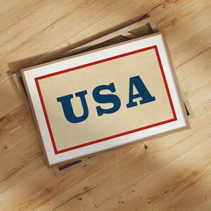 Puede incluir: Impresi&oacute;n enmarcada con la palabra "USA" en letras azules en negrita sobre un fondo beige, enmarcada con un borde rojo y un borde interior blanco. El marco es de color marr&oacute;n claro. La impresi&oacute;n est&aacute; sobre una superficie de madera.