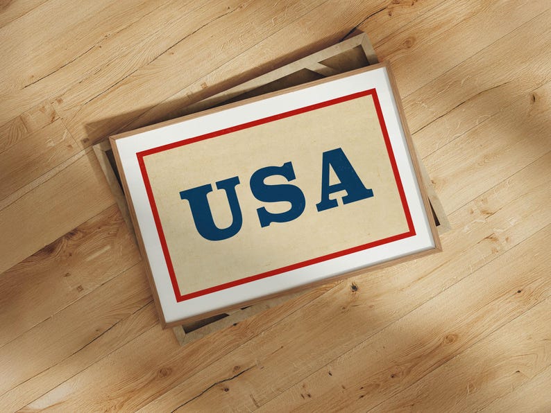 Puede incluir: Impresi&oacute;n enmarcada con la palabra "USA" en letras azules en negrita sobre un fondo beige, enmarcada con un borde rojo y un borde interior blanco. El marco es de color marr&oacute;n claro. La impresi&oacute;n est&aacute; sobre una superficie de madera.
