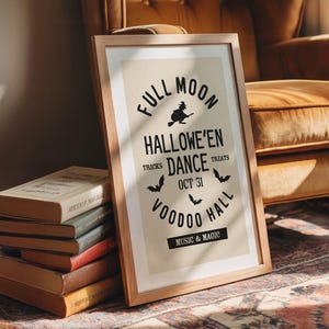 May include: A framed Halloween poster with a wooden frame. The poster features the text "Full Moon Hallowe'en Dance" with bats and a witch graphic. A stack of vintage books is in front of the poster.