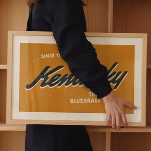 May include: A person holds a framed print with a yellow background and black text that reads "Kentucky, The Bluegrass State, Since 1792".