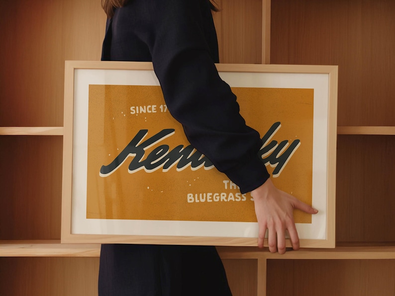 May include: A person holds a framed print with a yellow background and black text that reads "Kentucky, The Bluegrass State, Since 1792".