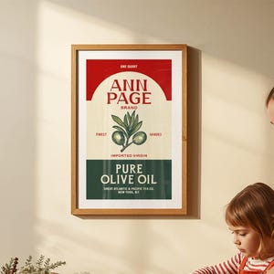 May include: A framed vintage-style print featuring "ANN PAGE BRAND" olive oil. The design includes a red and cream color scheme with the text "PURE OLIVE OIL" and an illustration of olive branches. The print is in a wooden frame.