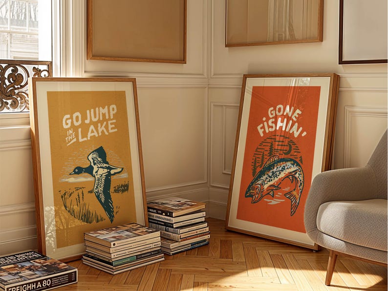 May include: Two framed prints with vintage-inspired designs. The left print features a yellow background with a duck in flight and the text "GO JUMP IN THE LAKE". The right print features an orange background with a fish jumping out of the water and the text "GONE FISHIN'".