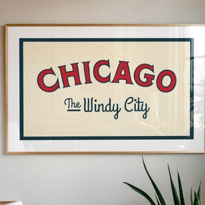 Op de afbeelding: Een ingelijste kunstdruk met het woord "CHICAGO" in rode, gebogen letters met een blauwe rand, boven "The Windy City" in kleinere, donkerblauwe letters. Het kunstwerk staat tegen een cr&egrave;mekleurige achtergrond, is goudkleurig omlijst en aan een witte muur bevestigd.