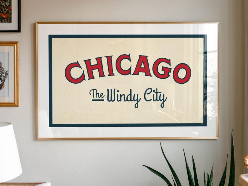 Op de afbeelding: Een ingelijste kunstdruk met het woord "CHICAGO" in rode, gebogen letters met een blauwe rand, boven "The Windy City" in kleinere, donkerblauwe letters. Het kunstwerk staat tegen een cr&egrave;mekleurige achtergrond, is goudkleurig omlijst en aan een witte muur bevestigd.