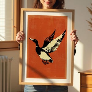 May include: A framed print of a stylized illustration of a duck with black and white markings flying against a burnt orange background.