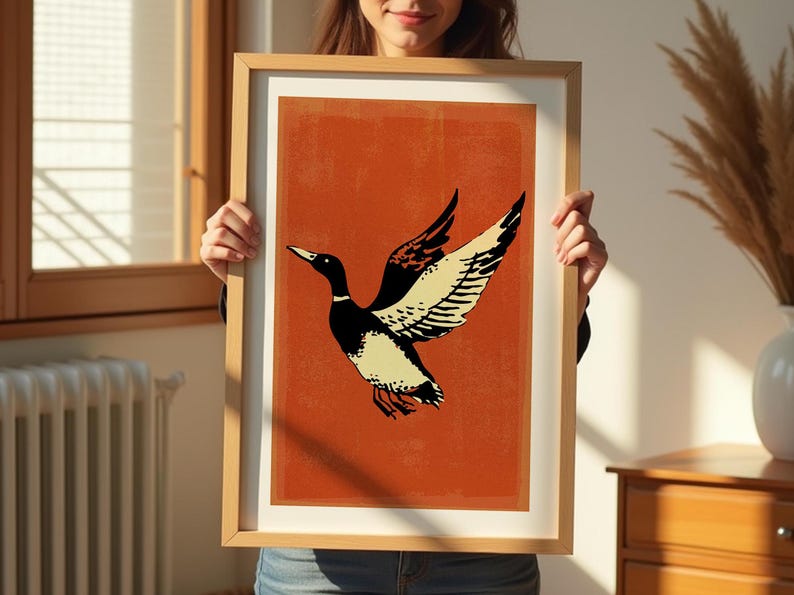 May include: A framed print of a stylized illustration of a duck with black and white markings flying against a burnt orange background.