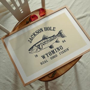 May include: A framed print featuring a detailed illustration of a fish, with text that reads "Jackson Hole Wyoming Real Good Fishin'". The print has a cream background and a wooden frame.