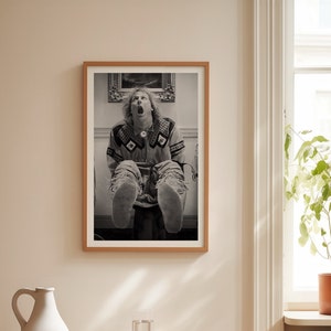 Op de afbeelding: Zwart-wit ingelijste print van een persoon met open mond en uitgestrekte benen. De persoon draagt een shirt met patroon en een gescheurde jeans. De print zit in een houten lijst en hangt aan een beige muur.