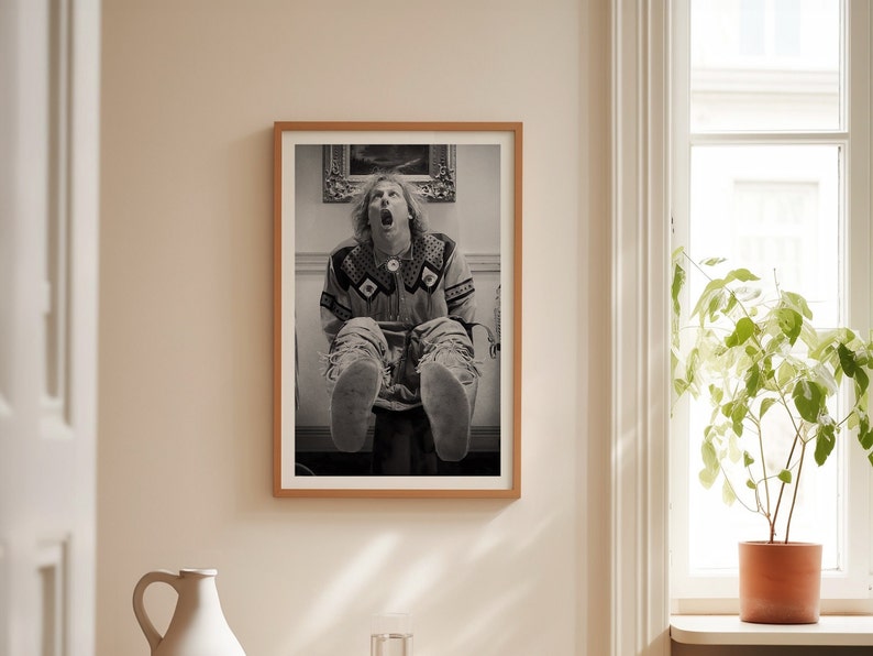 Op de afbeelding: Zwart-wit ingelijste print van een persoon met open mond en uitgestrekte benen. De persoon draagt een shirt met patroon en een gescheurde jeans. De print zit in een houten lijst en hangt aan een beige muur.