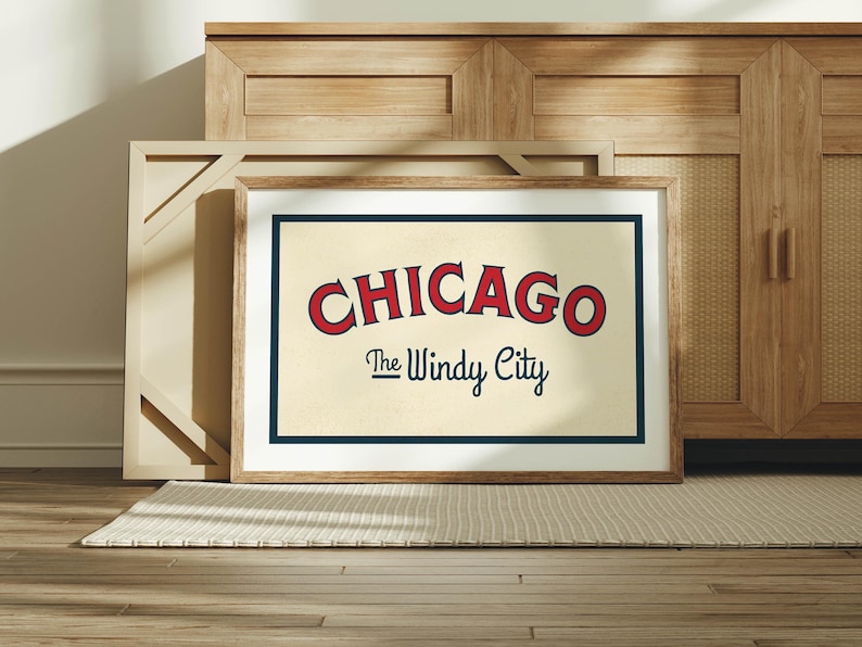 Op de afbeelding: Ingelijste kunstdruk met het woord "CHICAGO" in rode, gebogen letters boven "The Windy City" in een scriptlettertype. Het kunstwerk heeft een cr&egrave;mekleurige achtergrond met een marineblauwe rand en een houten lijst. De print wordt op een tapijt weergegeven.