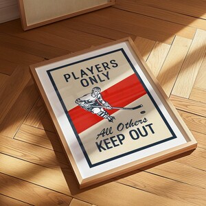 Pu&ograve; includere: Stampa artistica incorniciata a tema hockey. L'opera presenta un'illustrazione di un giocatore di hockey con una striscia diagonale rossa e il testo "Players Only, All Others Keep Out". La stampa &egrave; in una cornice di legno chiaro.