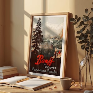 Op de afbeelding: Een ingelijste vintage reisposter voor Banff in de Canadese Rockies, met een berglandschap met een stad en een grote dennenboom. De poster heeft de tekst "Banff" in rood script en "Canadian Pacific". Een koffiekopje en een open boek liggen op tafel.