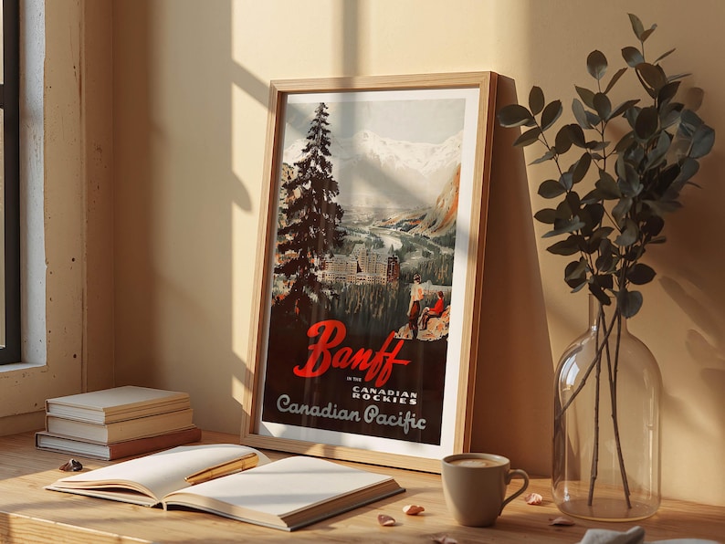 Op de afbeelding: Een ingelijste vintage reisposter voor Banff in de Canadese Rockies, met een berglandschap met een stad en een grote dennenboom. De poster heeft de tekst "Banff" in rood script en "Canadian Pacific". Een koffiekopje en een open boek liggen op tafel.