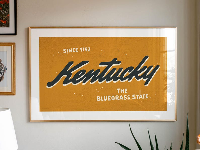 May include: A framed print with a gold frame and a yellow background. The print features the text "Kentucky, The Bluegrass State" in black lettering with the words "Since 1792" above the state name.