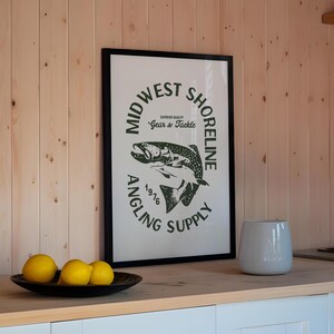 May include: A framed print with a black frame and a white background. The print features a green logo with the text "Midwest Shoreline Angling Supply 1976" and a graphic of a fish. The print is hanging on a wall with wood paneling.