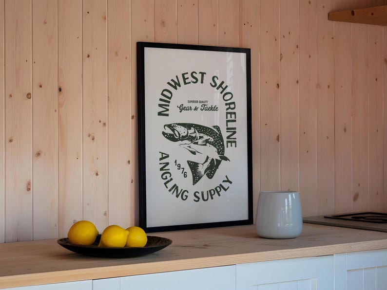May include: A framed print with a black frame and a white background. The print features a green logo with the text "Midwest Shoreline Angling Supply 1976" and a graphic of a fish. The print is hanging on a wall with wood paneling.