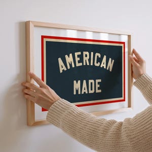 May include: A framed print with the words "AMERICAN MADE" in a vintage style. The text is cream-colored against a dark blue background, framed by a red border and a natural wood frame. The print is being held up against a white wall.
