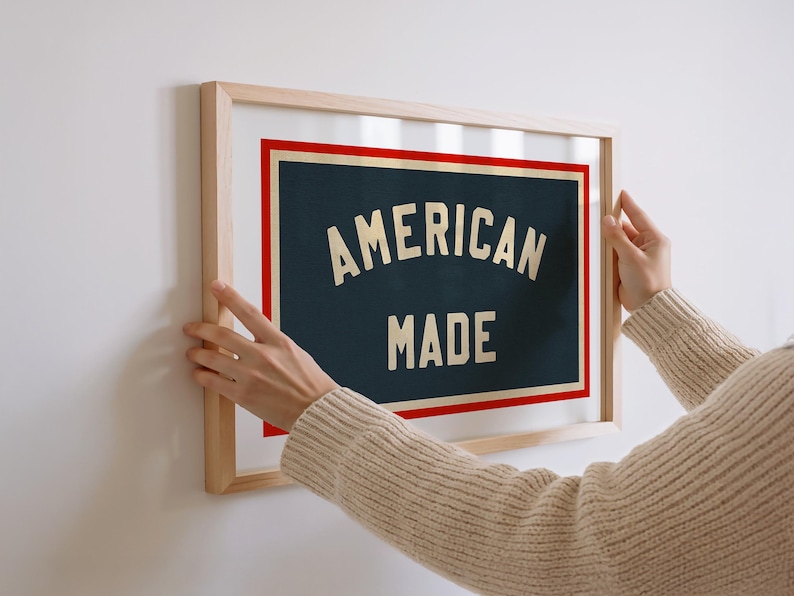 May include: A framed print with the words "AMERICAN MADE" in a vintage style. The text is cream-colored against a dark blue background, framed by a red border and a natural wood frame. The print is being held up against a white wall.