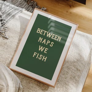 May include: A framed art print with the text "BETWEEN NAPS WE FISH" in a serif font. The artwork features a dark green background with the text in a light beige color. The frame is a light brown color. The print is resting on a light-colored, textured surface.