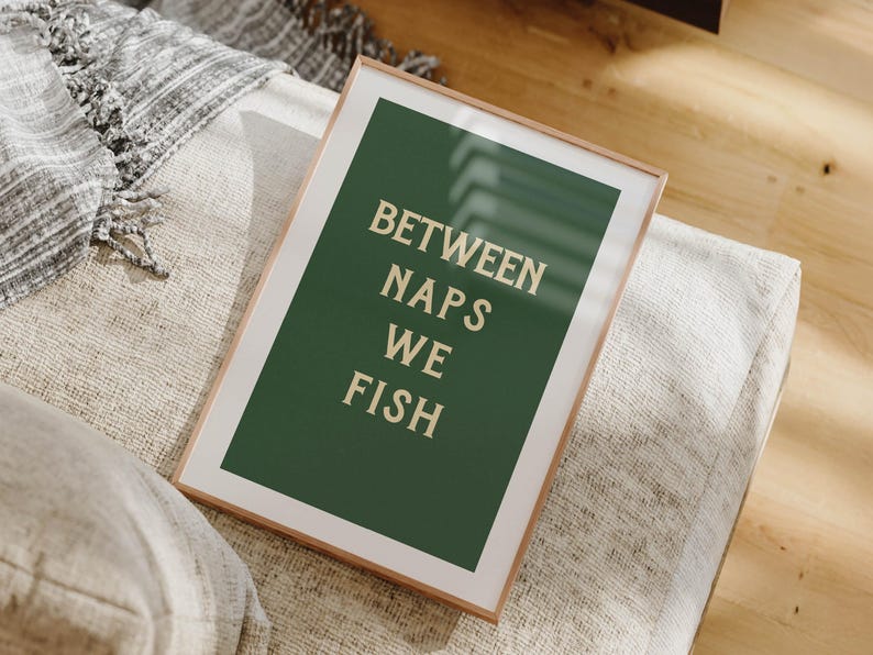 May include: A framed art print with the text "BETWEEN NAPS WE FISH" in a serif font. The artwork features a dark green background with the text in a light beige color. The frame is a light brown color. The print is resting on a light-colored, textured surface.