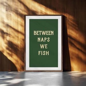 May include: Framed art print with the text "BETWEEN NAPS WE FISH" in a serif font on a dark green background. The print is in a brown frame and is leaning against a wooden wall. The wall has a natural wood grain.