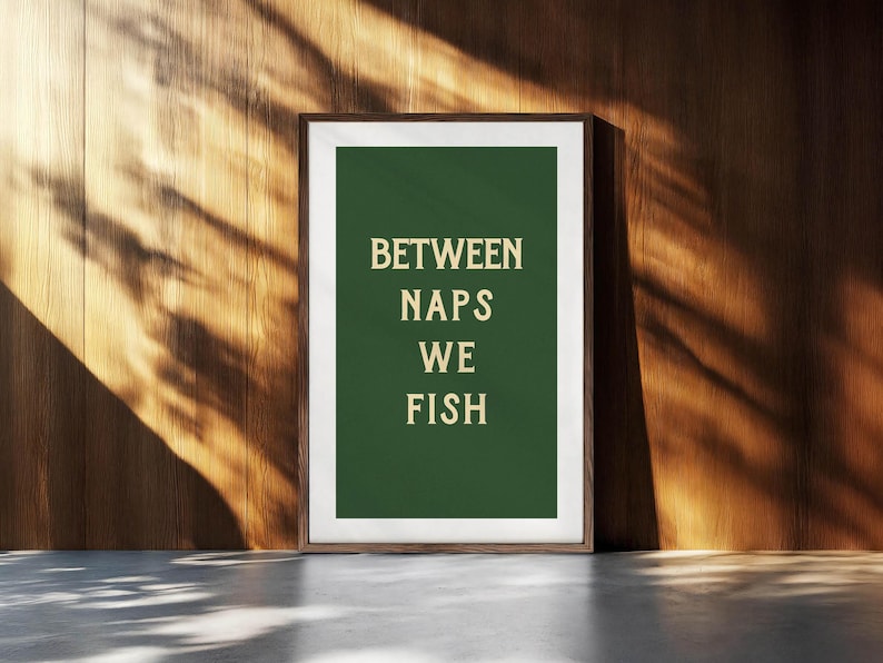 May include: Framed art print with the text "BETWEEN NAPS WE FISH" in a serif font on a dark green background. The print is in a brown frame and is leaning against a wooden wall. The wall has a natural wood grain.
