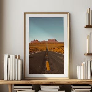 May include: A photograph of a long, straight road leading towards a mountain range in the distance. The road is paved and has a yellow dividing line. The mountains are brown and rocky. The sky is blue and clear.