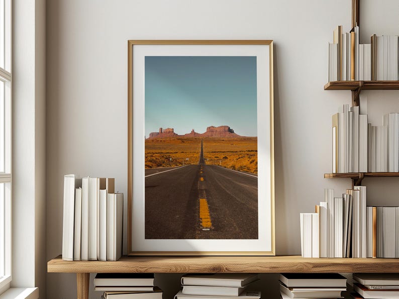 May include: A photograph of a long, straight road leading towards a mountain range in the distance. The road is paved and has a yellow dividing line. The mountains are brown and rocky. The sky is blue and clear.