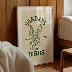 May include: A framed poster with a light beige background and green text and illustration. The text reads "SUNDAYS ARE FOR THE BIRDS" with an illustration of an eagle. The poster is displayed in a room with wooden paneling.
