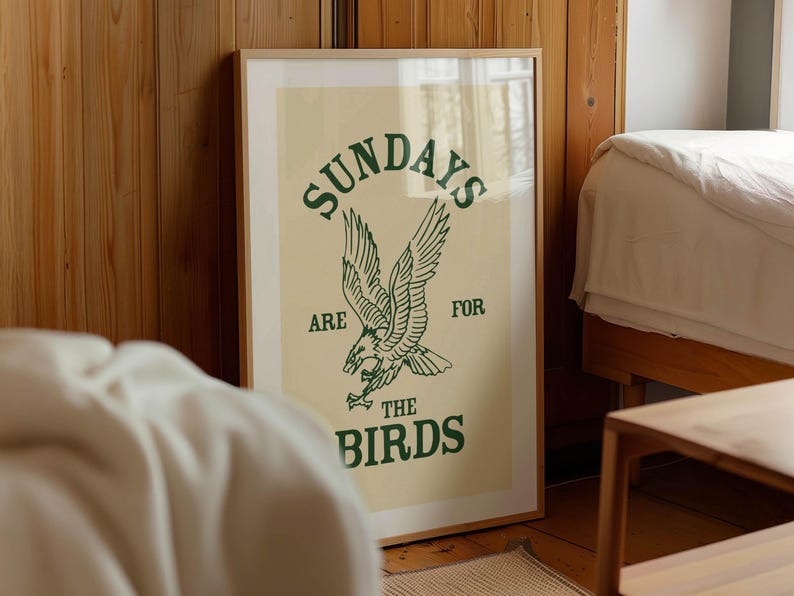 May include: A framed poster with a light beige background and green text and illustration. The text reads "SUNDAYS ARE FOR THE BIRDS" with an illustration of an eagle. The poster is displayed in a room with wooden paneling.