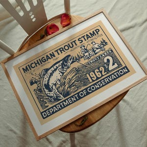May include: Framed vintage print of a Michigan Trout Stamp from 1962. The artwork features a leaping trout, fishermen, and the text "Department of Conservation." The print is in a light wood frame.