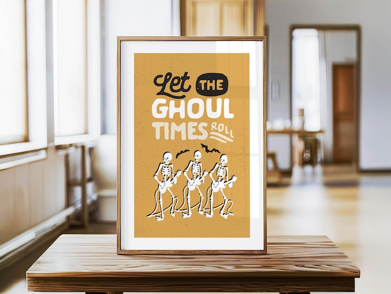May include: Framed art print with a mustard yellow background featuring the text "Let the Ghoul Times Roll." Three skeleton figures play guitars below the text. The artwork is in a wooden frame and sits on a wooden table.