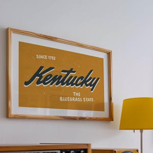May include: A framed print with a yellow background and black text that reads "Kentucky, The Bluegrass State, Since 1792".