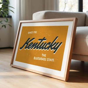 May include: A framed print with a yellow background and black text that reads "Kentucky, The Bluegrass State, Since 1792".